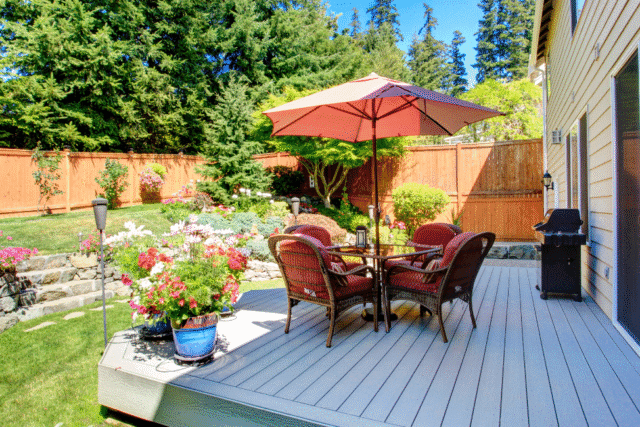 Sunny backyard with a raised deck featuring a patio table, red cushioned chairs, and a large umbrella, surrounded by lush landscaping, colorful flowers, and a wooden privacy fence
