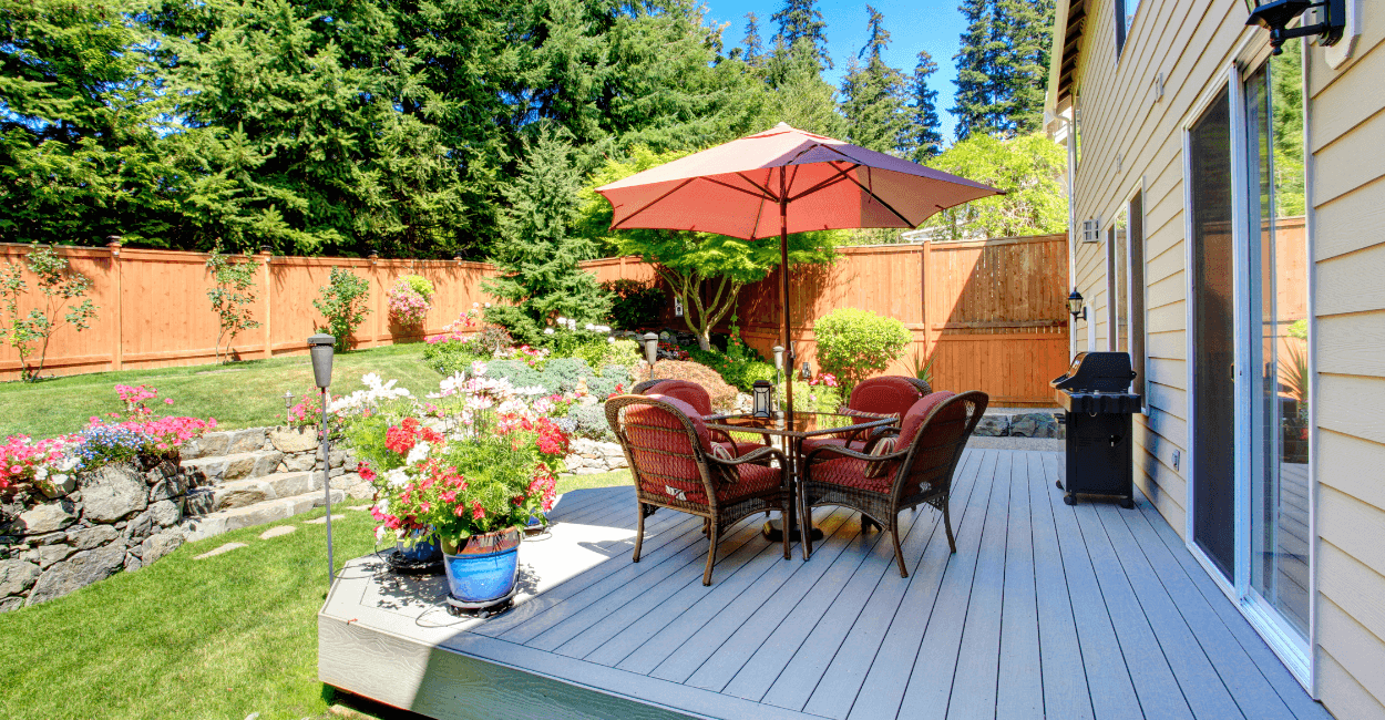 Sunny backyard with a raised deck featuring a patio table, red cushioned chairs, and a large umbrella, surrounded by lush landscaping, colorful flowers, and a wooden privacy fence